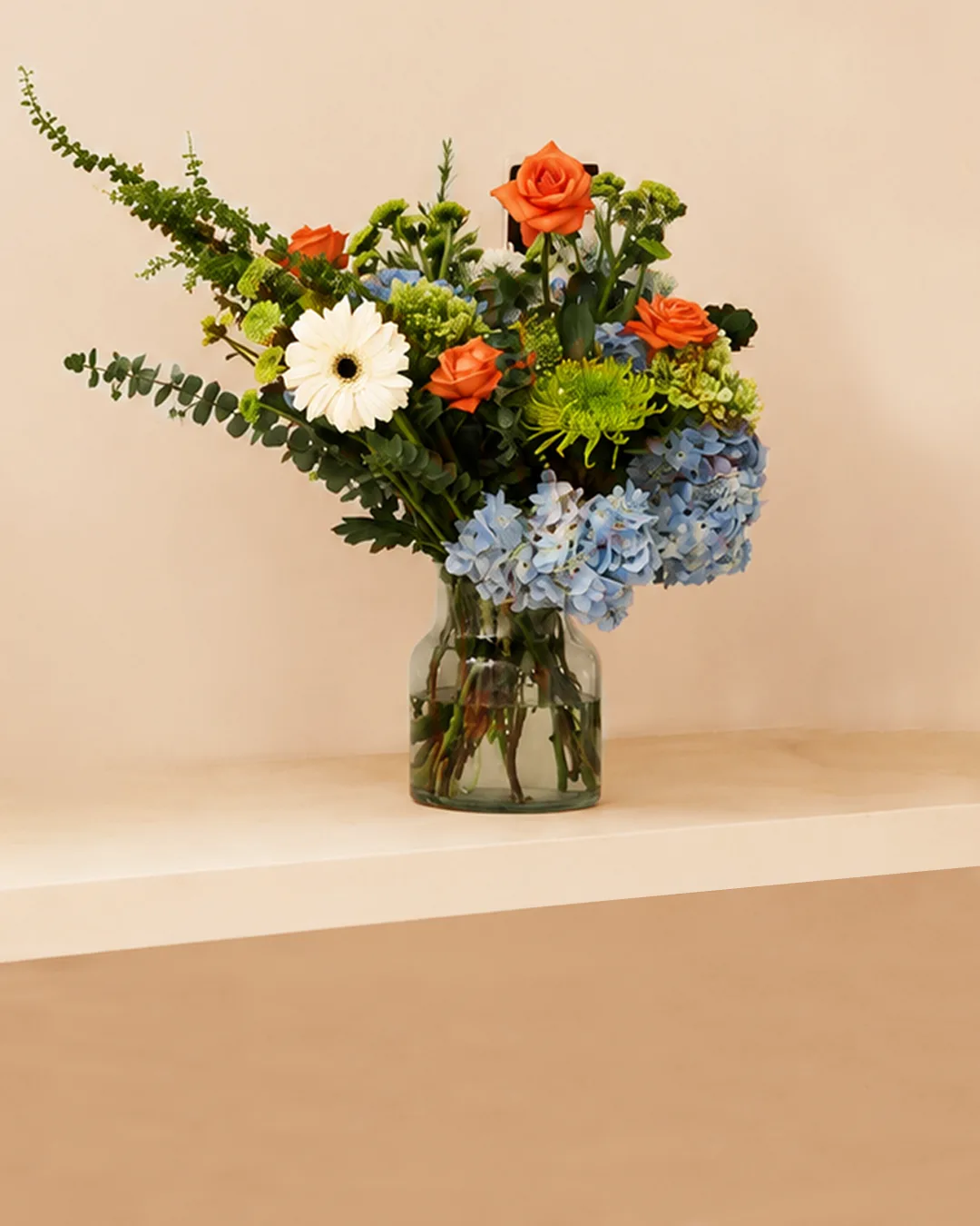 Blue hydrangea and orange rose arrangement in a glass vase against a beige wall